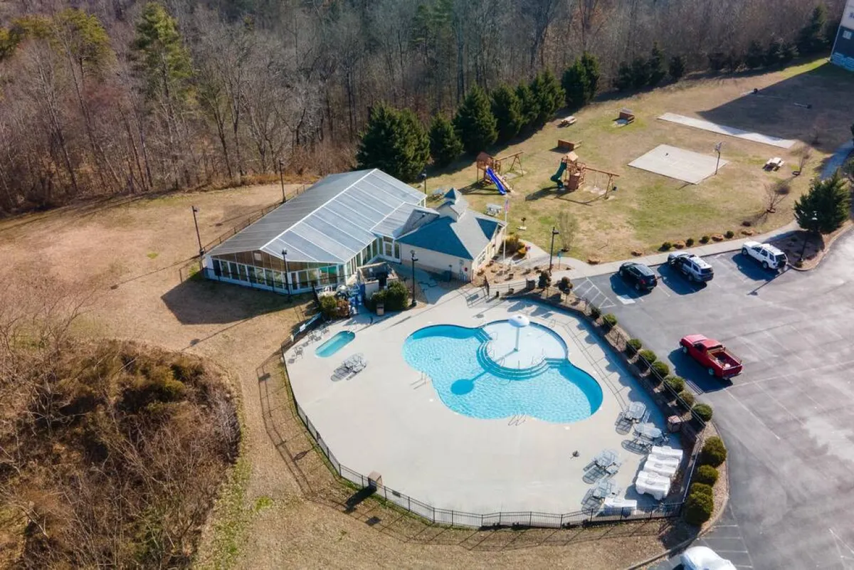This lively exterior scene presents the outdoor swimming pool area, surrounded by lush greenery, with recreational spaces and playground equipment visible. Children can be seen playing, while adults relax by the pool, evoking a sense of cheerful family fun under the open sky.