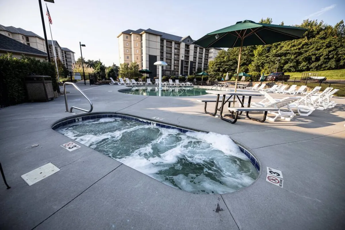 An inviting outdoor pool area featuring a sparkling swimming pool, a bubbling hot tub, lounge chairs for sunbathing, a picnic table, and a large umbrella. The setting is lively and family-friendly, emphasizing leisure and social gatherings.