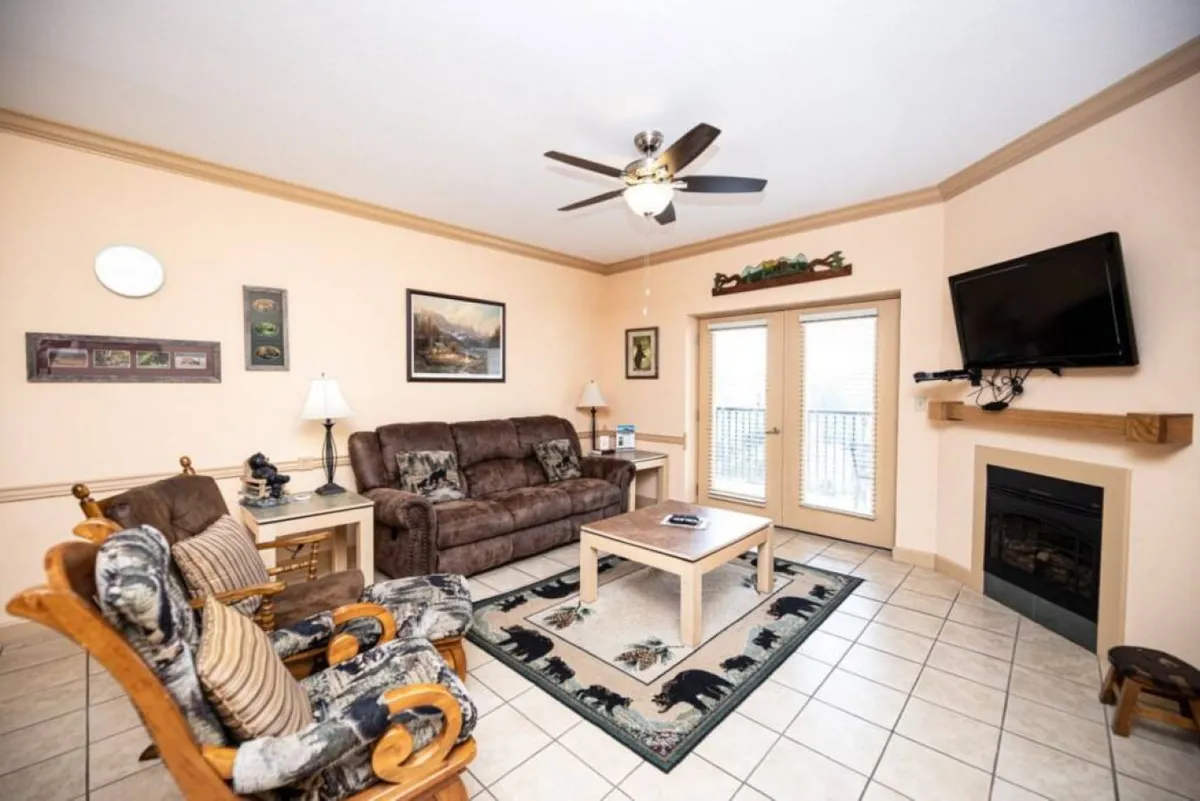 A spacious living area featuring a comfortable sofa, coffee table, fireplace, and a flat-screen television. Large viewing windows fill the room with natural light and offer glimpses of the surrounding mountain landscape. The setup invites relaxation and socializing, making it ideal for movie nights or enjoying morning coffee while taking in the view.