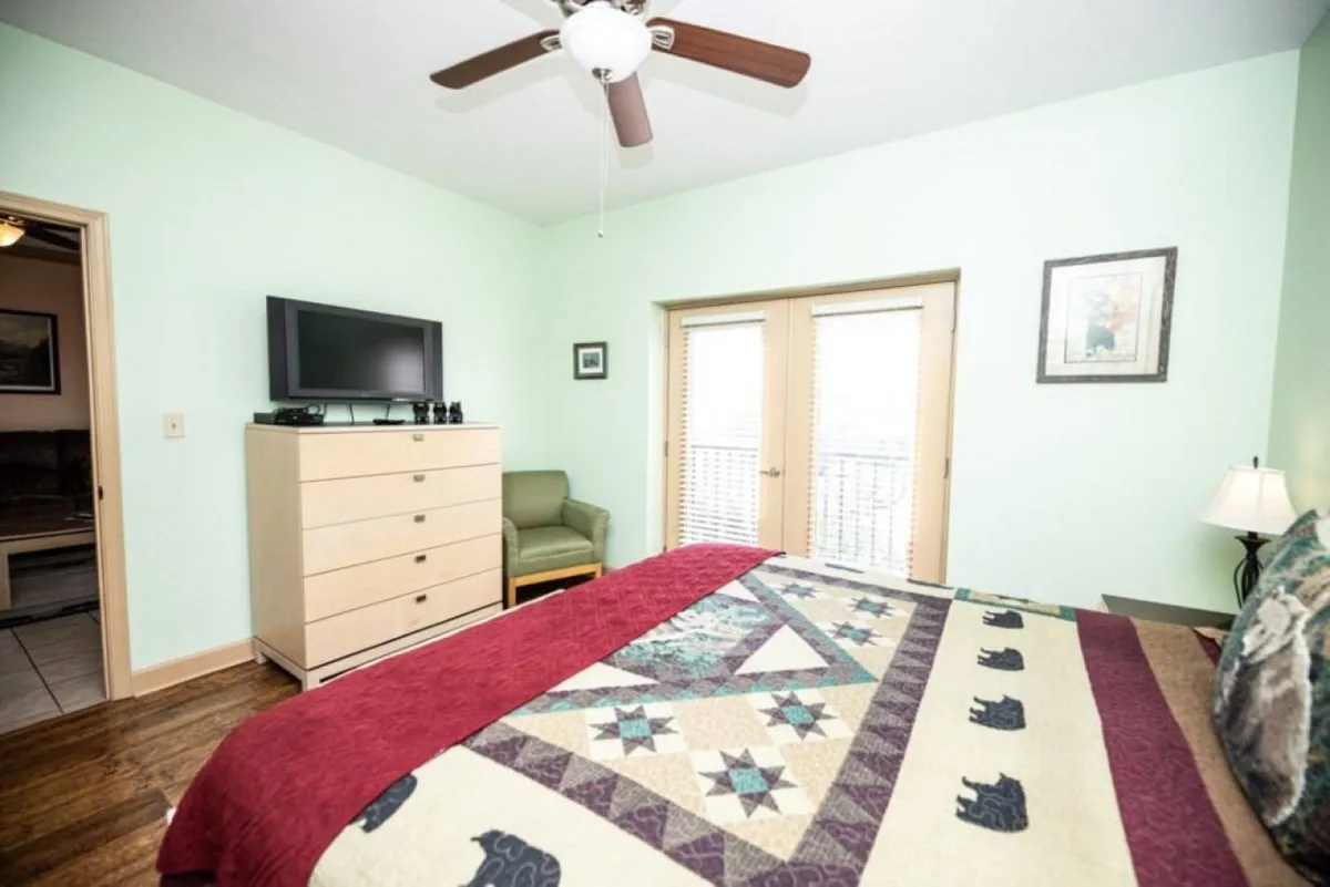 A spacious bedroom showcasing a bedspread with bear motifs, a wooden dresser with a television, and balcony doors letting in natural light. The layout highlights a comfortable king-size bed and a serene, inviting atmosphere.