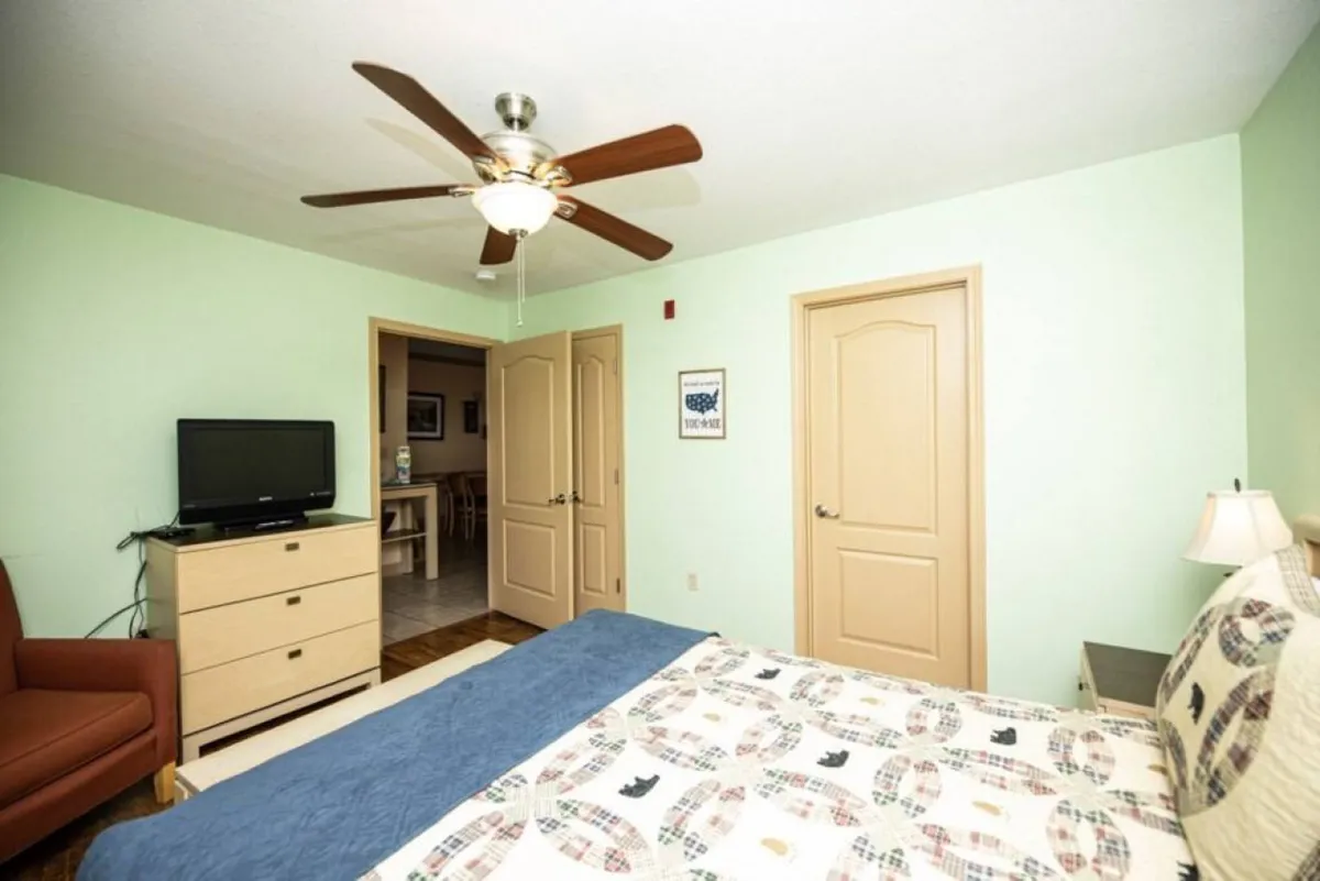 A cozy bedroom with a queen-size bed, patterned comforter, ceiling fan, a TV atop a dresser, and an armchair by the window, all arranged for optimal comfort and relaxation.