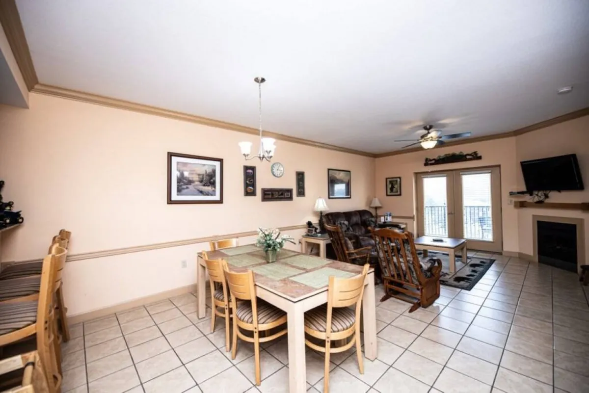 An open-plan space featuring a dining table, reclining chair, fireplace, and sliding doors leading to a balcony. Wall art and warm decor enhance the cozy feel. The setup encourages gathering for meals, enjoying the fireplace, or accessing the balcony to soak in the mountain views.