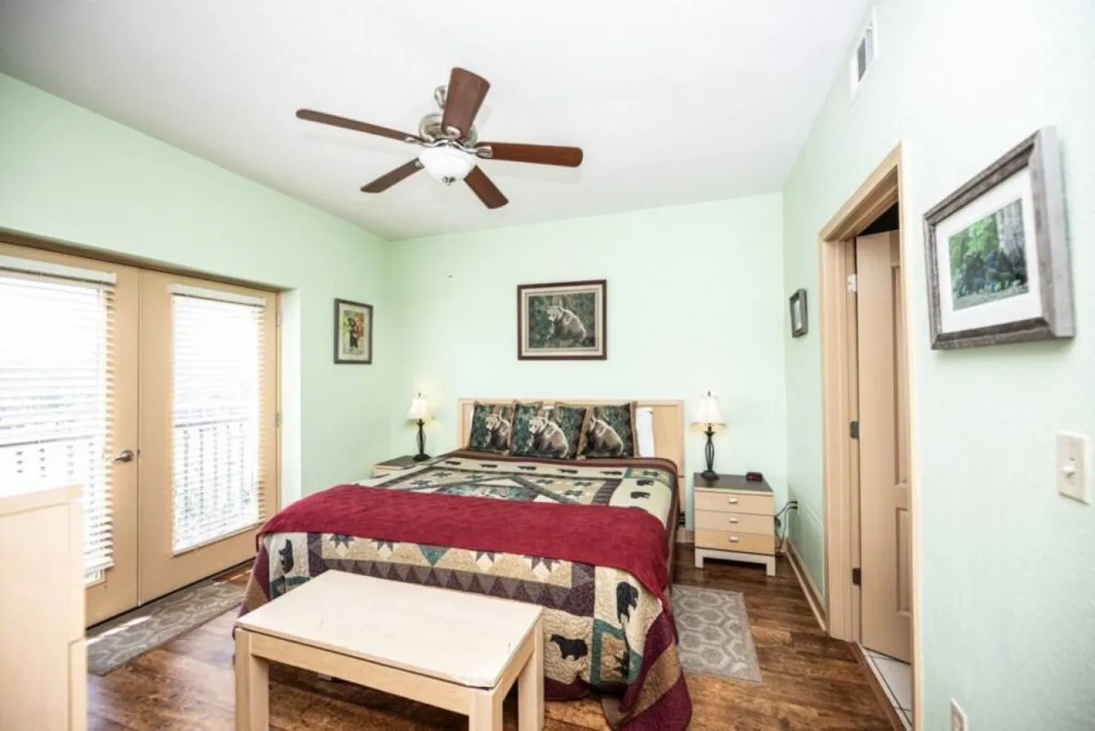 The image shows a warm and inviting bedroom interior with a plush, quilted bed as the centerpiece, flanked by wooden nightstands. Sunlight streams through large windows with blinds, casting a natural glow across the space. Tasteful artwork brightens the walls, creating a tranquil and welcoming atmosphere.