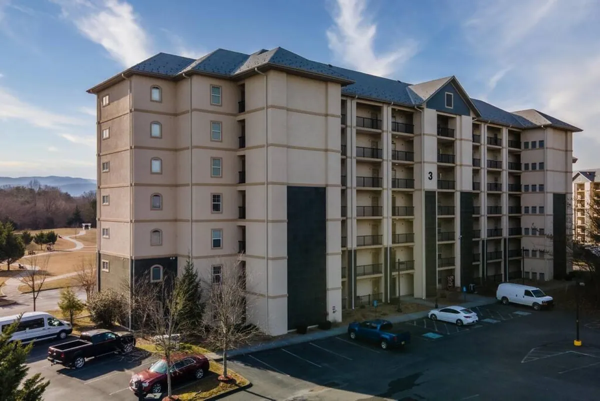 A welcoming exterior shot of The Cove's five-story building, surrounded by landscaped parking and set against a scenic mountain backdrop. The image captures the property's inviting façade, ample parking, and its beautiful natural surroundings.